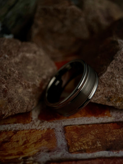 A brushed dome-shaped tungsten ring with a polished groove, placed on a textured surface.