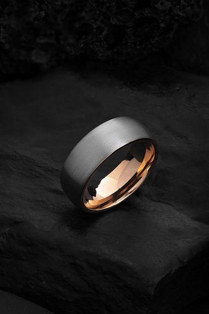 A grey tungsten wedding band with gold detailing, displayed on a dark, textured surface.