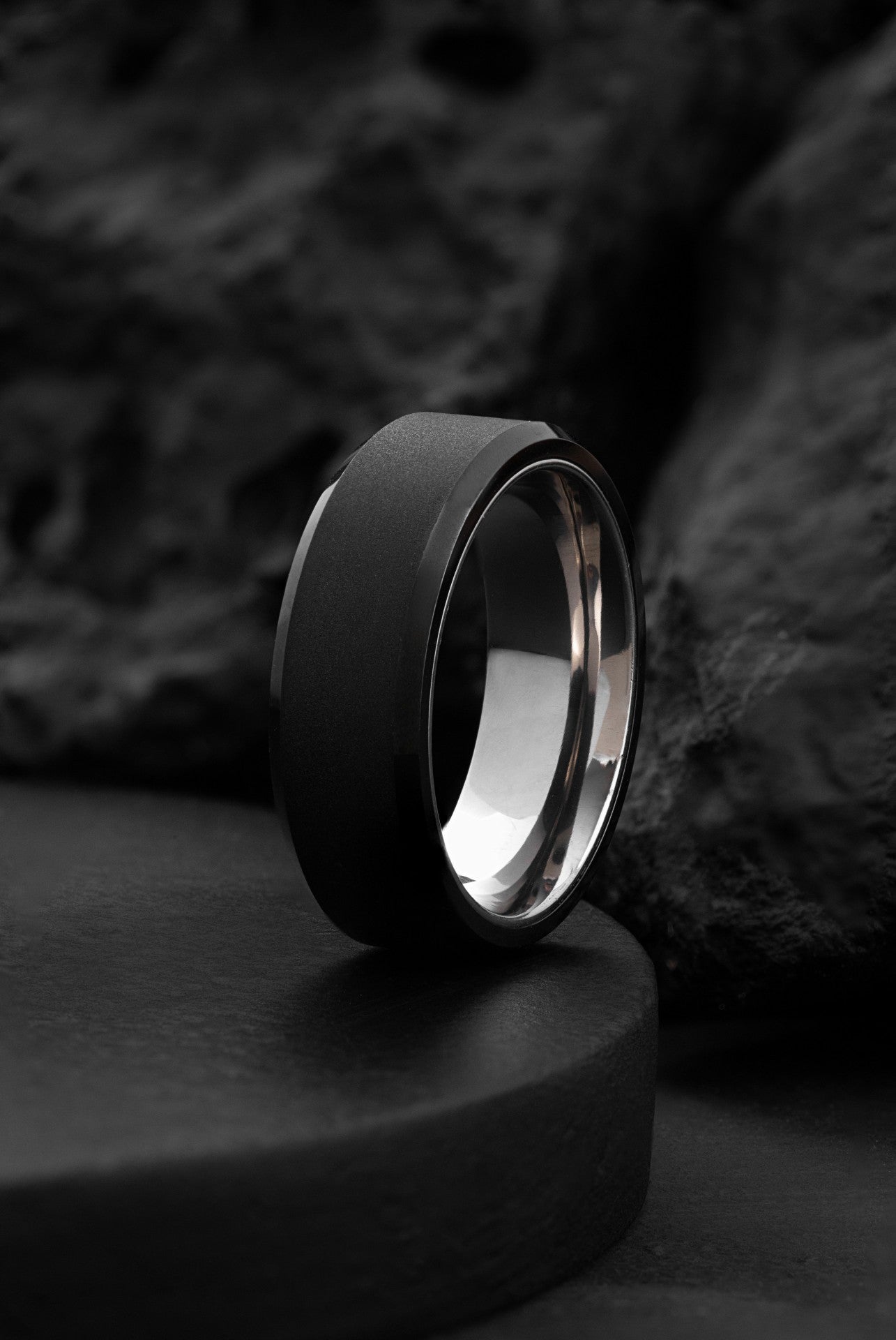 A contemporary titanium and black brushed tungsten ring with beveled edges, placed on a dark surface.