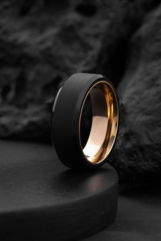 A black tungsten carbide ring with a rose gold band, placed on a dark surface, showcasing its bold and minimalist design.