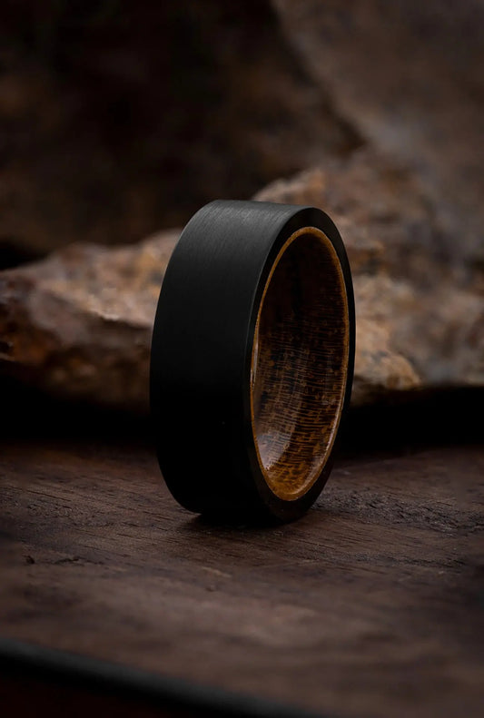 A black tungsten band ring with a red wood patterned top, set on a textured dark background.