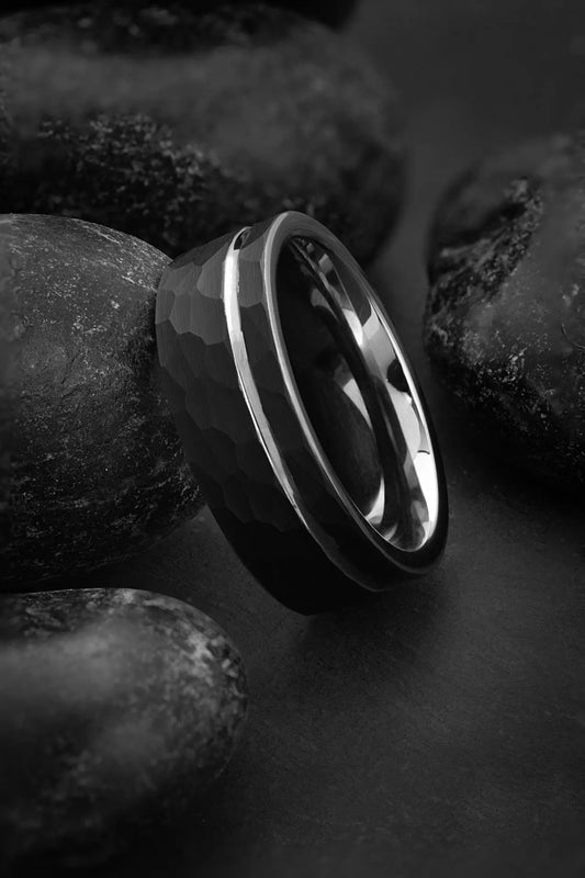 A black tungsten carbide band ring with a polished silver interior and an offset grey inlay, displayed on a textured dark background.