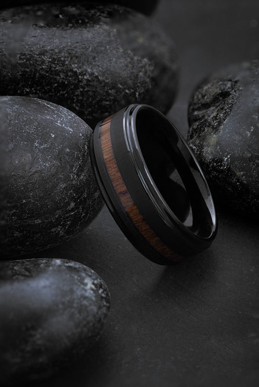A Tungsten carbide and Hawaiian Koa wood ring with a black band and a wood inlay, placed on a textured dark background, showcasing its design and material.