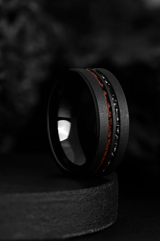 A black wedding band ring with two inlays, one featuring a red-colored edge and the other a dark, textured surface, set against a blurred background.