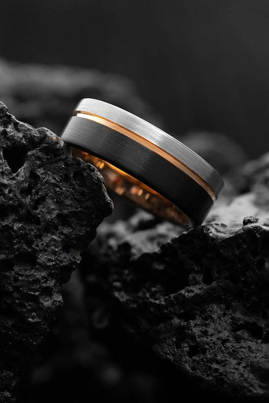 A wedding band with a gray tungsten carbide exterior and a rose gold inlay, resting on a textured black surface.