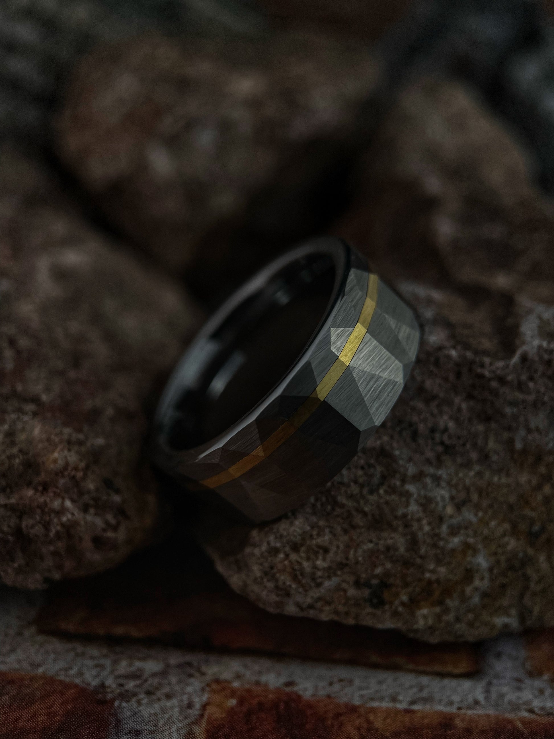 A tungsten carbide and 14k gold ring with a distinctive faceted texture and gold inlay, displayed on a rocky surface.