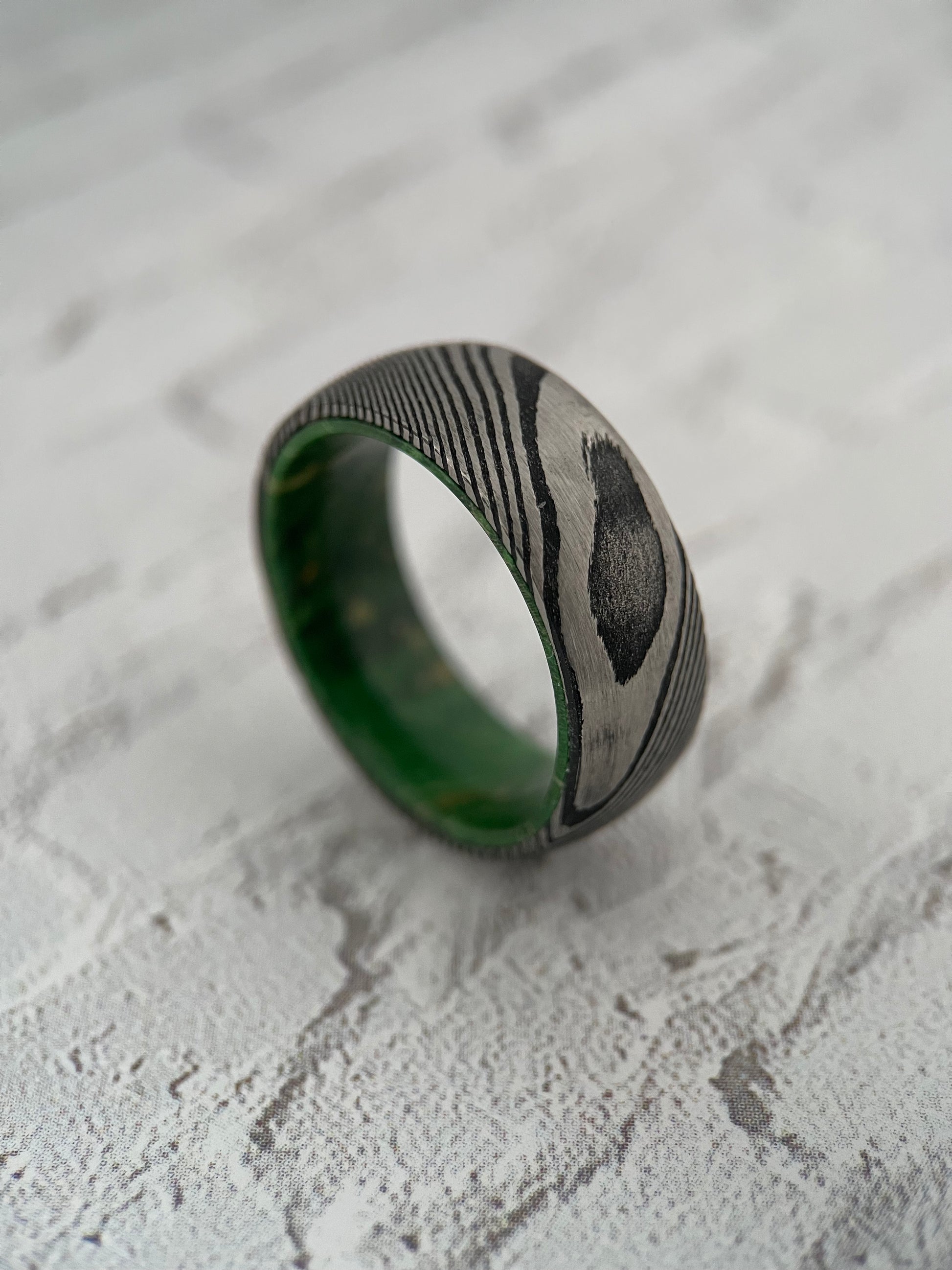 The Iris Damascus steel ring featuring a vibrant green alder wood interior sleeve.