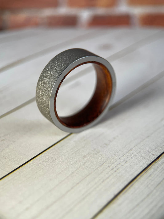 Close-up of Regalia Rings' The Prometheus Redwood Liner and Sandblasted Tungsten Band, showcasing its textured surface and rich wood-tone inlay.