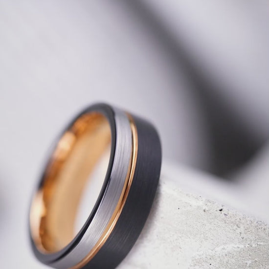 A wedding band ring with a distinctive desert Ironwood interior and a brushed matte Tungsten exterior featuring a rose gold inlay, resting on a textured black surface.