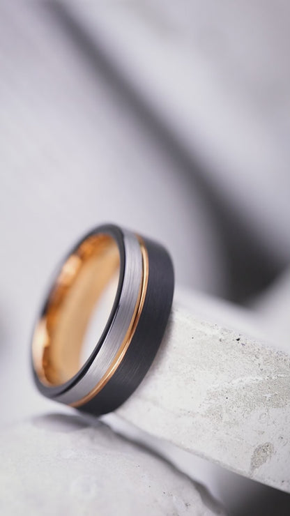A wedding band ring with a distinctive desert Ironwood interior and a brushed matte Tungsten exterior featuring a rose gold inlay, resting on a textured black surface.
